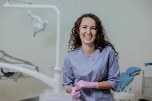 Dental assistant in a patient treatment room smiling at the camera and wearing pink gloves.