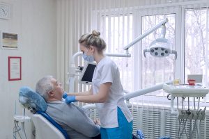 Dentist assistant in protective mask with elderly patient before examination in dental office.