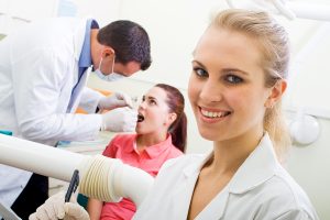 Young happy dental assistant in a dentist practice with a dentist and patient in the background.