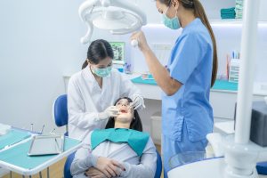 Female dentist adjust dental surgical light then starts checking or examining tooth of young girl patient lying on dental chair. Dental assistant supports procedure by handing instruments to dentist.