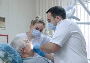 Male dentist with nurse in protective masks working with patient in clinic. Senior man getting treatment at dental office. Dentist and assistant during teamwork. Healthcare medicine concept
