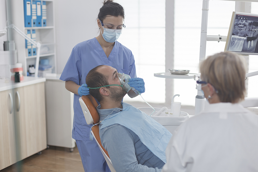 Man sitting in dentists chair with dentist sitting next to him and dental assistant standing on the opposite side