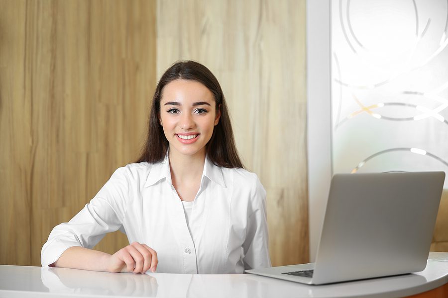 Young female receptionist sitting at dental office front desk in front of a laptop smiling