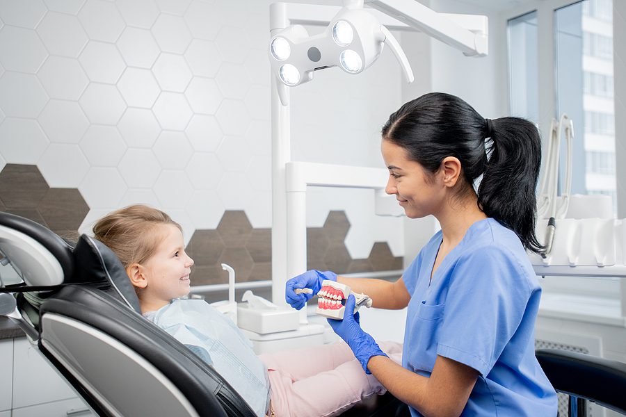 Dental assistant in blue uniform showing young patient in armchair how to brush teeth properly while sitting in front of her.