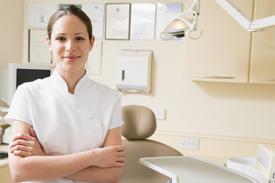 Female dental assistant in white scrubs smiling at the camera with her arms folded. 