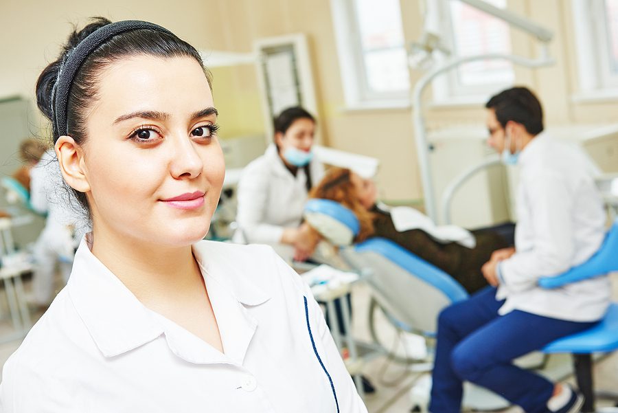 A female dental assisting student smiles confidently in front of a dentist and patient. 
