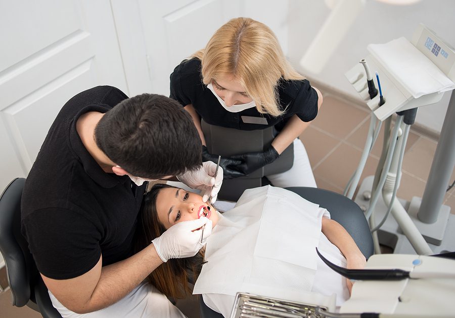 Male dentist with female dental assistant in training, leaning over patient observing. 