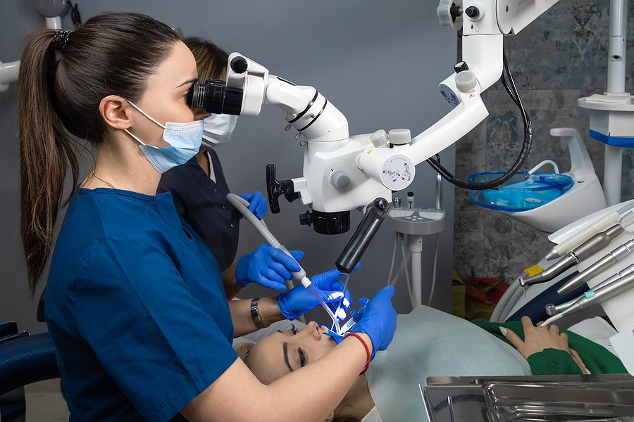 Dental assistant wearing blue scrubs is looking through a dental microscope. 