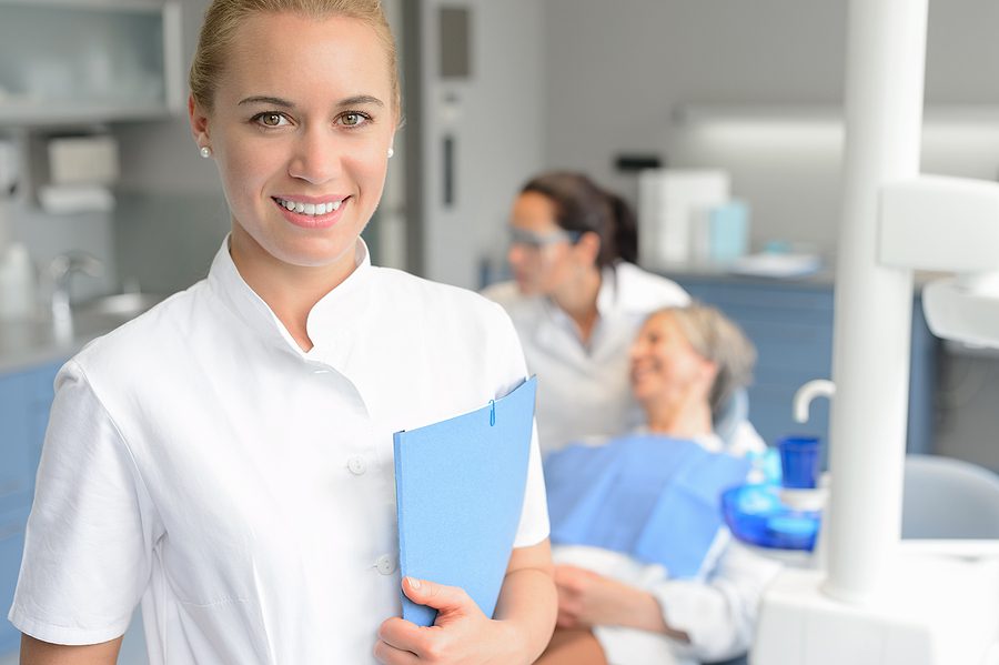 Female dental assistant holding a blue patient chart and smiling at camera with dentist and patient blurred in background. 