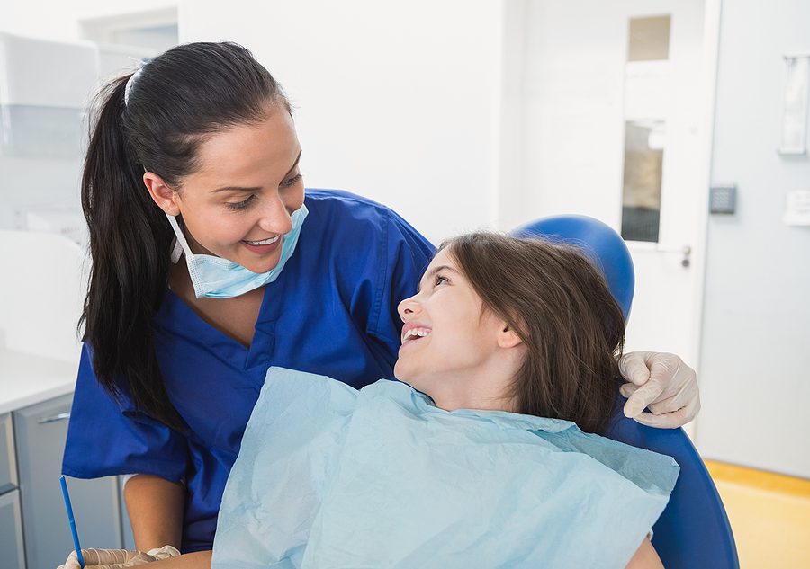 Female dental assistant wearing blue scrubs smiling at a pediatric patient in a dental clinic. 