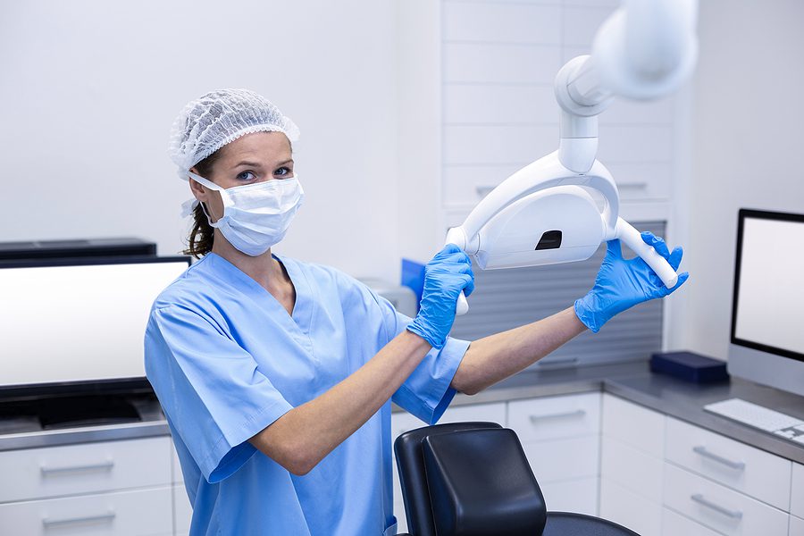 Female dental assistant wearing blue scrubs and a medical mask, adjusting a light in a dental exam room. 