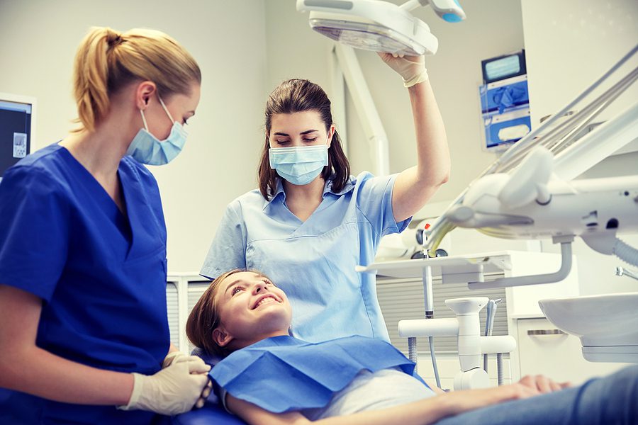 Female dentist and dental assistant wearing blue scrubs, performing a dental exam on a female pediatric patient. 