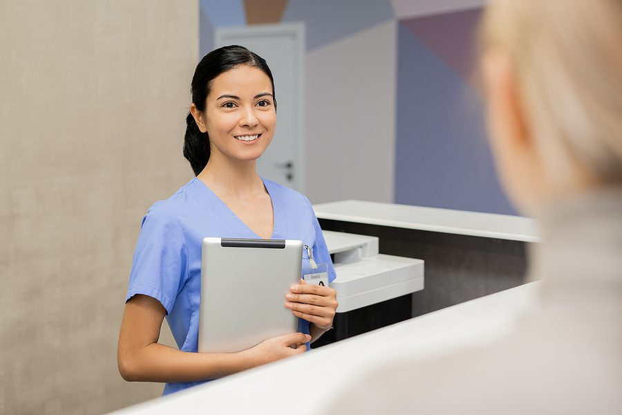 Female dental assistant wearing blue scrubs, holding a patient file and smiling at the camera. 