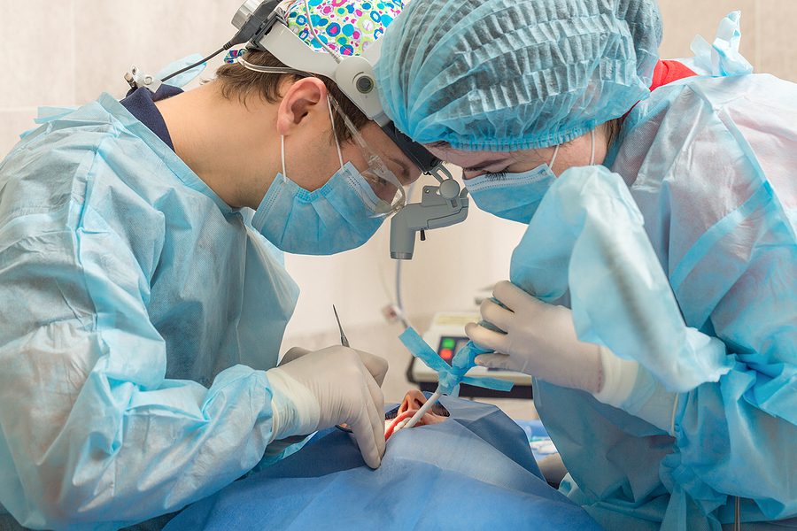 Dentist and dental assistant wearing surgical scrubs, performing oral surgery on a patient. 