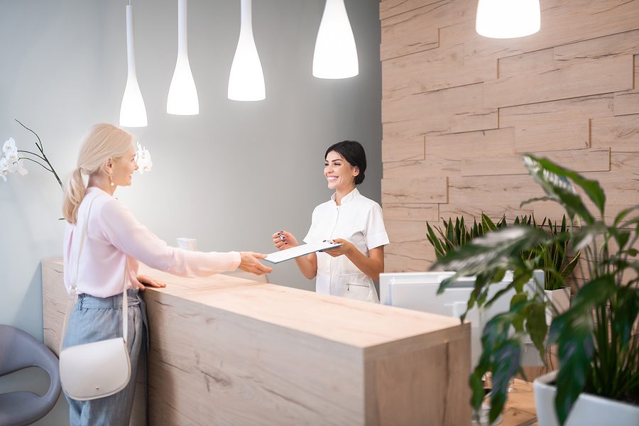 Woman standing at the reception desk of a dental clinic while the front desk administrator hands her a clipboard. 