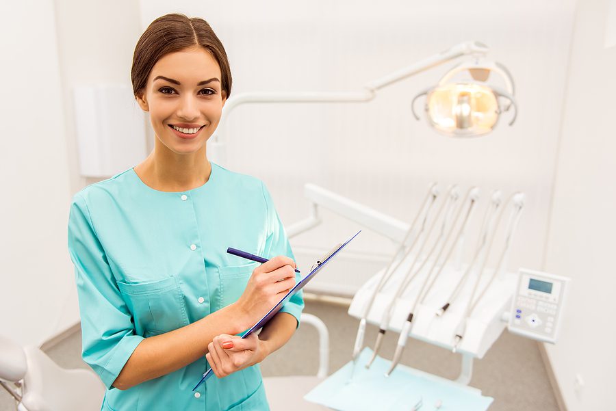 Female dental assistant wearing green scrubs, standing in front of a dental chair and holding a clipboard. 